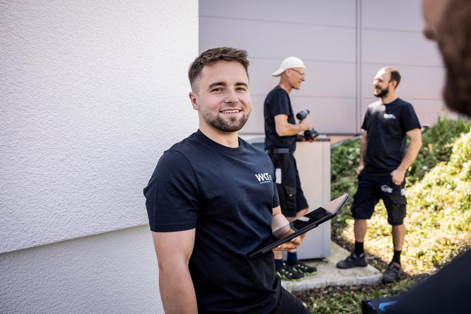 Mitarbeiter im WKT-Shirt lächelt mit Tablet in der Hand vor zwei Handwerkern an einer Wärmepumpe