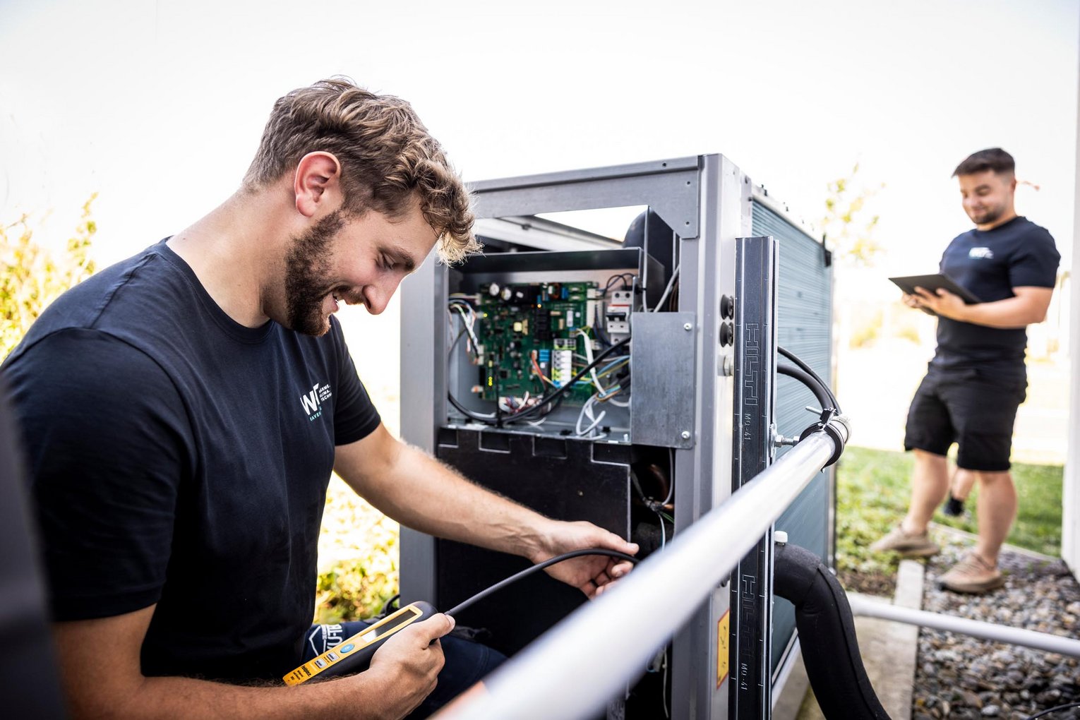 Techniker prüft Kabelverbindungen einer Außenwärmepumpe, während ein Kollege Notizen macht