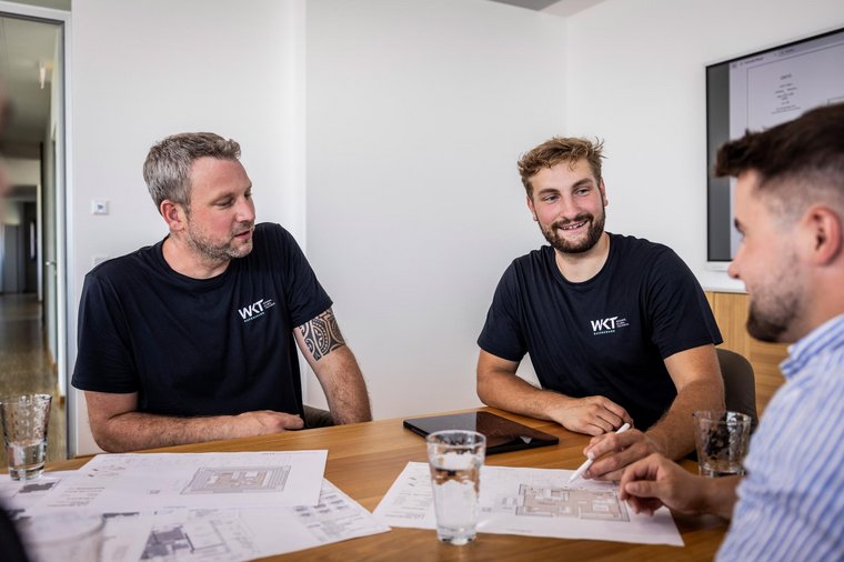 Zwei WKT-Mitarbeiter im schwarzen Shirt besprechen Baupläne mit Kunden an einem Holztisch